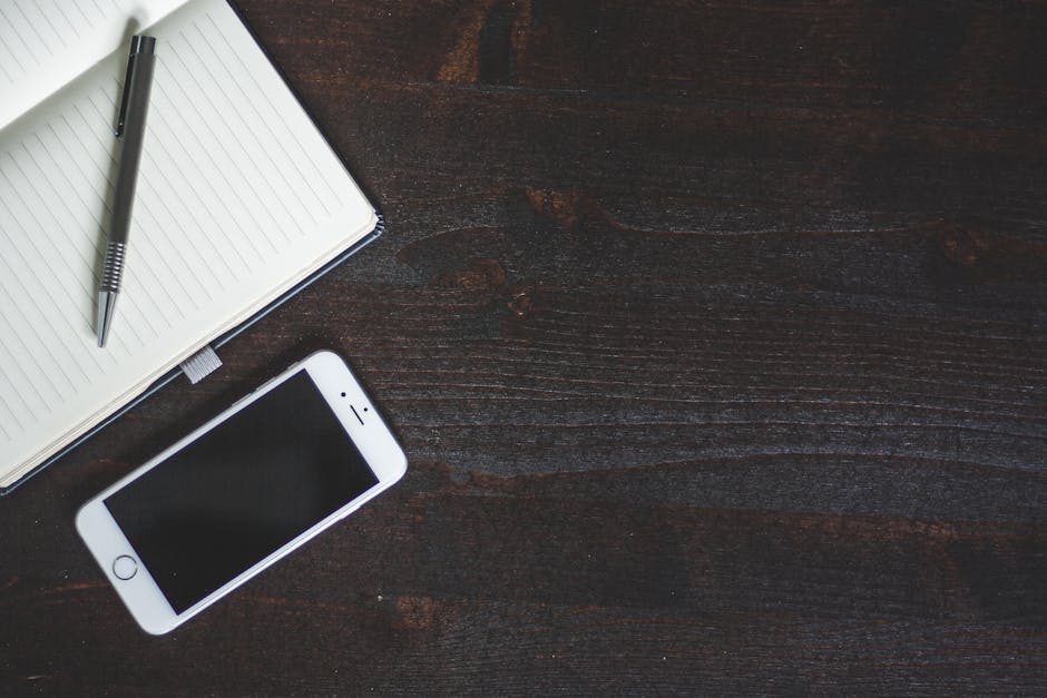 A clean and minimalist workspace featuring a smartphone, notebook, and pen on a dark wood table.